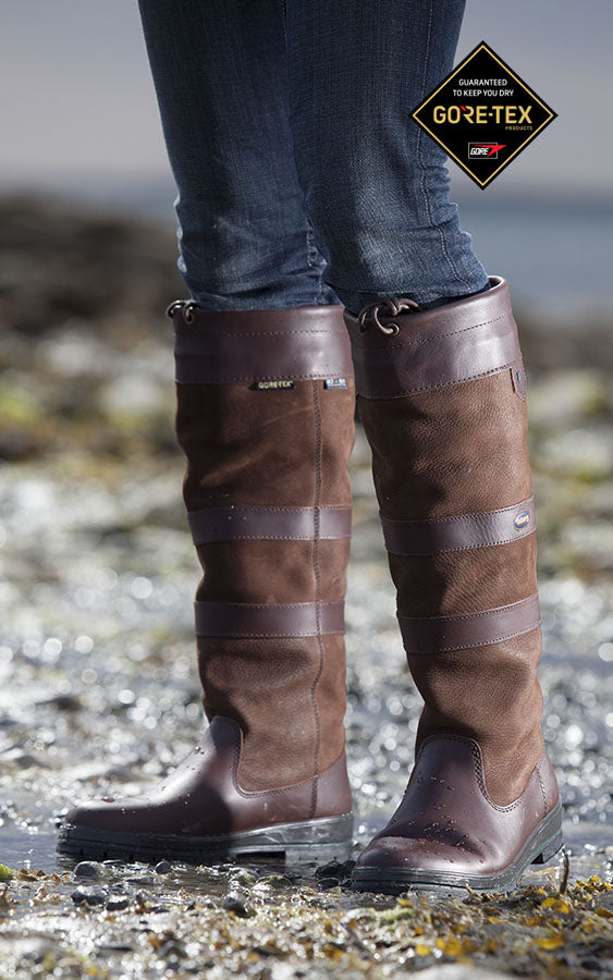 Brown waterproof knee-high boots with GORE-TEX logo worn by a person on a rocky surface.