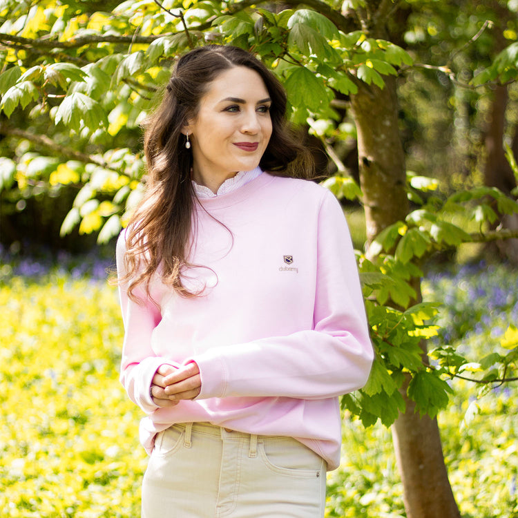 Square image of a woman wearing a pink sweatshirt from Dubarry of Ireland stands in a garden beside a tree on a sunny day.//© Jade Brook | @ townlifecountrystyle