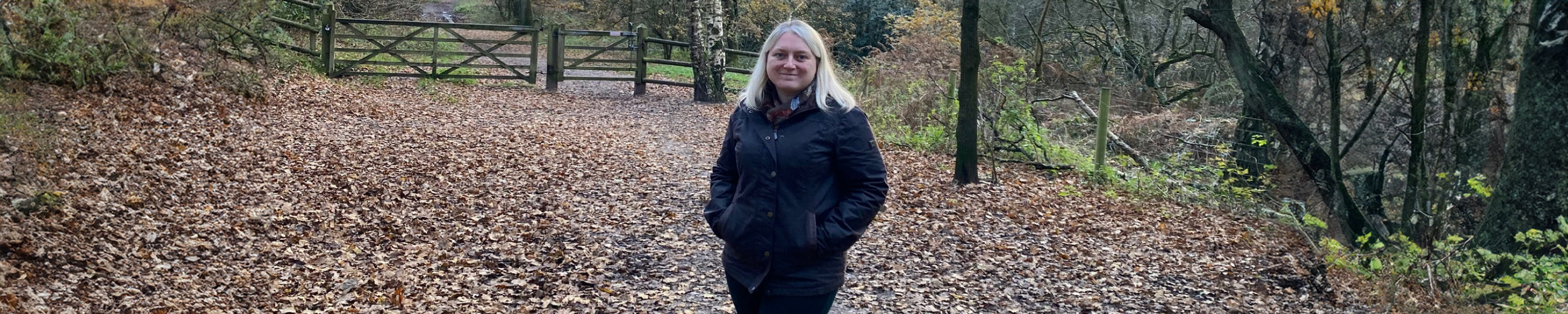 A wide image of a woman wearing a Mountrath Wax Jacket in a forest in an autumnal setting with brown leaves littering the floor.