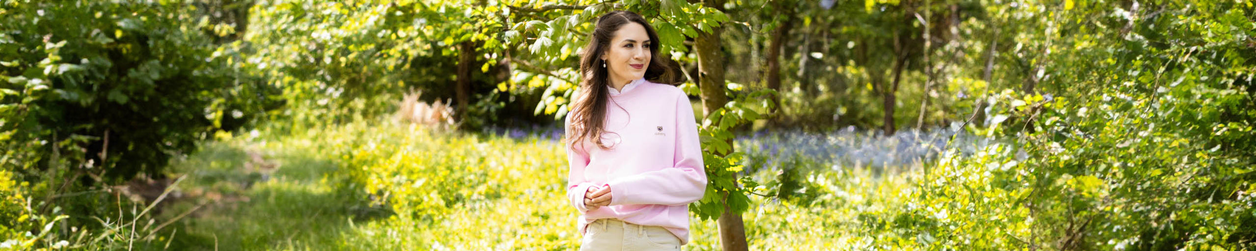 A wide image of a woman wearing a pink sweatshirt from Dubarry of Ireland stands in a garden beside a tree on a sunny day.