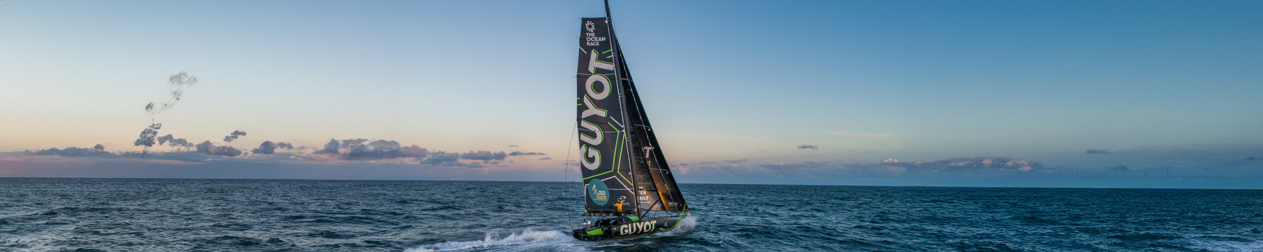 Wide image of Team Guyot's green and black boat out on the ocean during a beautiful sunset in the background.