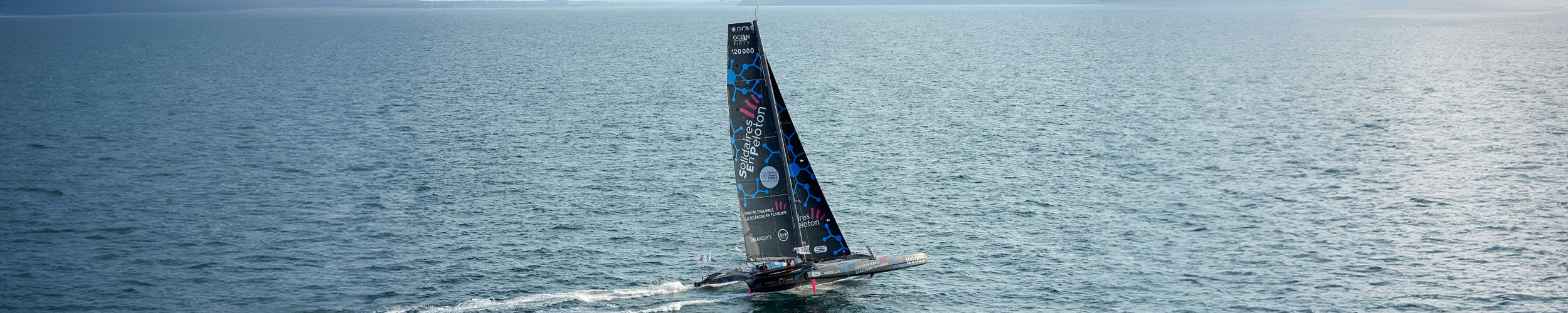 Wide image of Thibaut Vauchel-Camus' vessel sails on a sunny day with blue skies and a blue ocean.